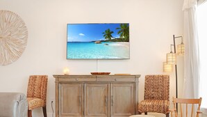 Un salon avec un grand téléviseur à écran plat fixé au mur, qui diffuse une scène de plage, un meuble en bois, deux fauteuils à motifs, un petit pouf et une lampe sur pied.