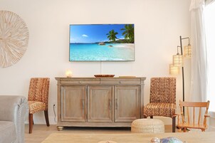 A living room with a large flat-screen TV mounted on the wall displaying a beach scene, a wooden cabinet, two patterned armchairs, a small ottoman, and a floor lamp.