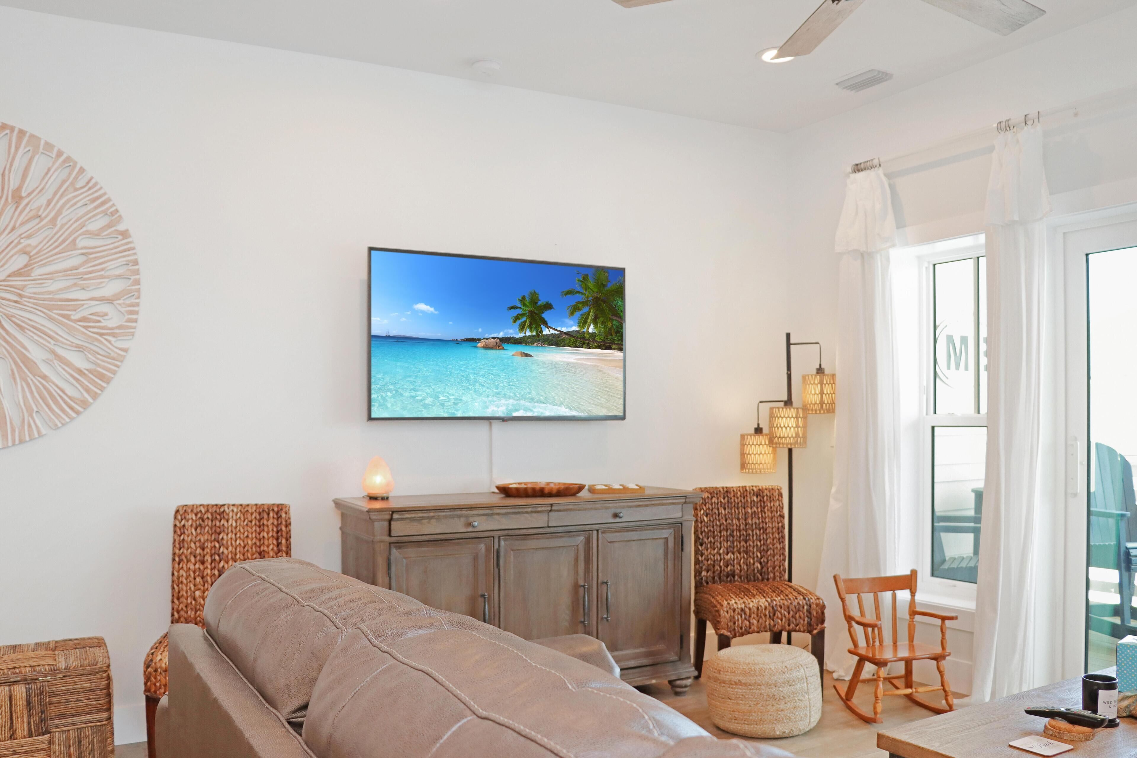Un salon avec un grand téléviseur à écran plat diffusant une scène de plage, un canapé, des fauteuils en osier, un buffet en bois et une table à manger avec une bougie.