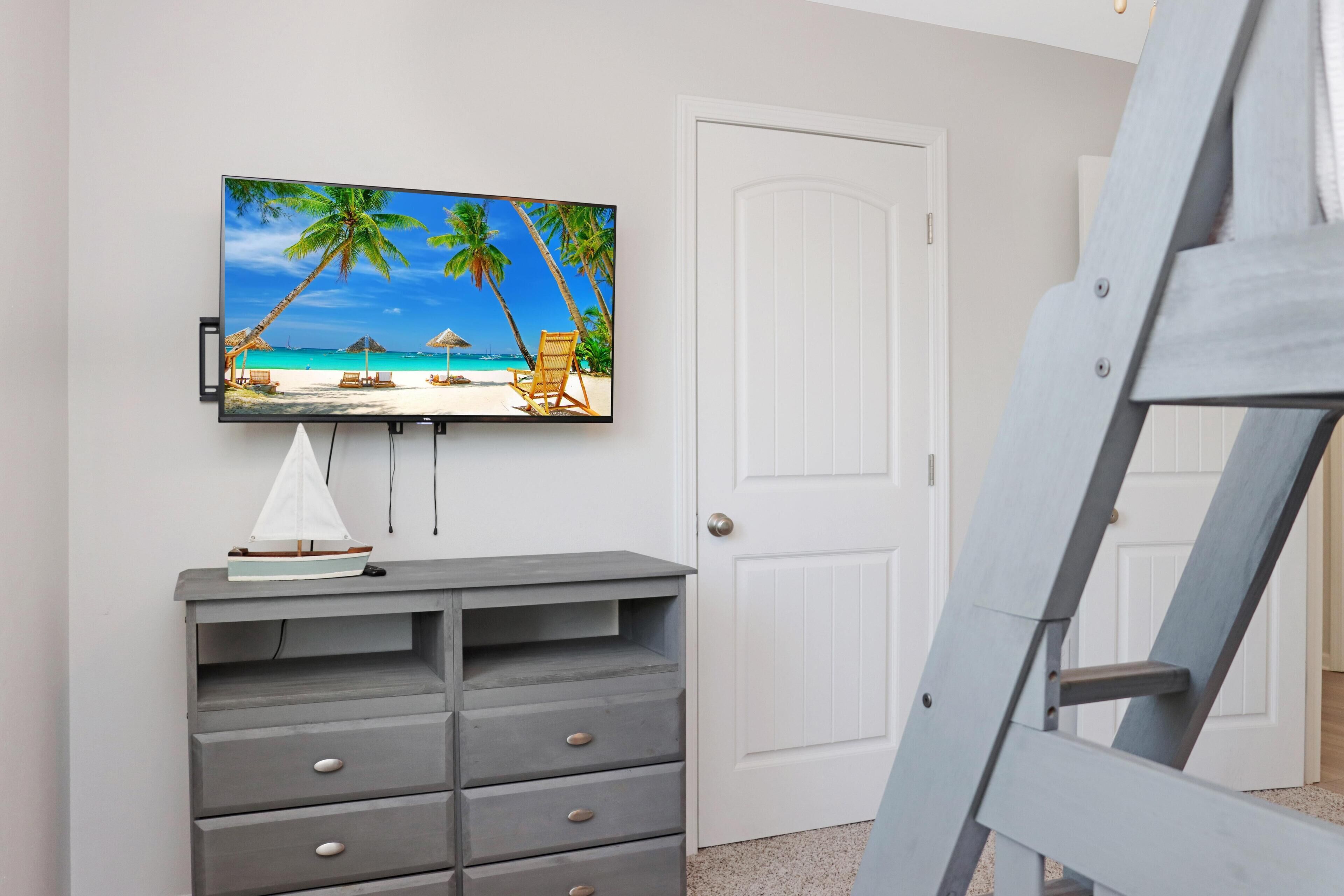 A room with a television mounted on the wall displaying a beach scene, a grey dresser with drawers, and a white door.