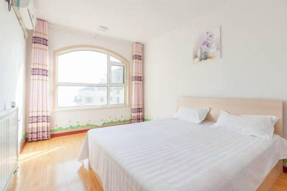 A bedroom with a large bed, a window with curtains, and a wall-mounted artwork.