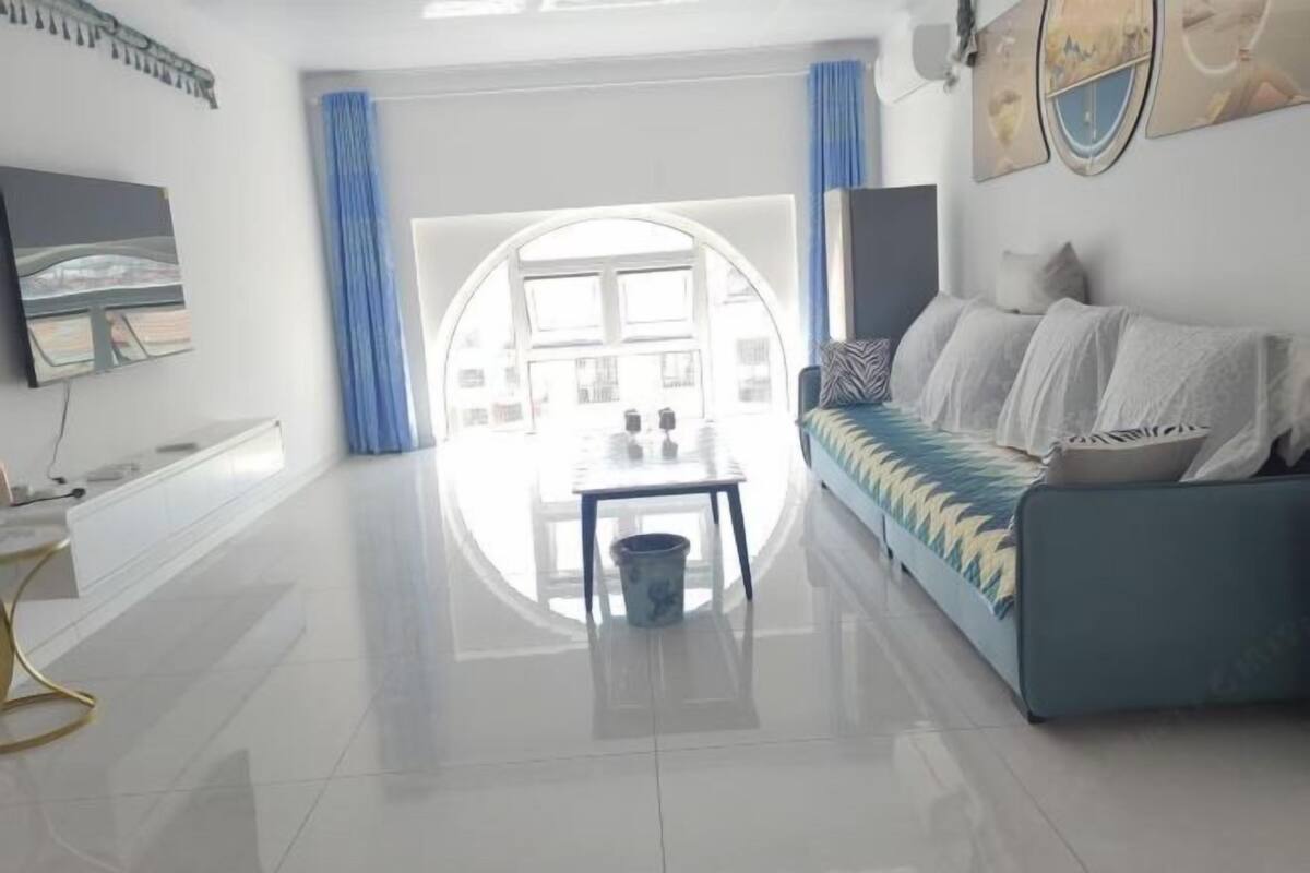 A modern living room with a sofa, a TV, a dining table, and a view through a large window.