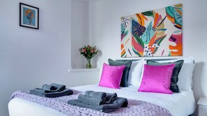 Un lit bien fait, recouvert d’une couverture violette, avec deux oreillers et des serviettes pliées. Un tableau abstrait aux couleurs vives est accroché au mur, au-dessus du lit.