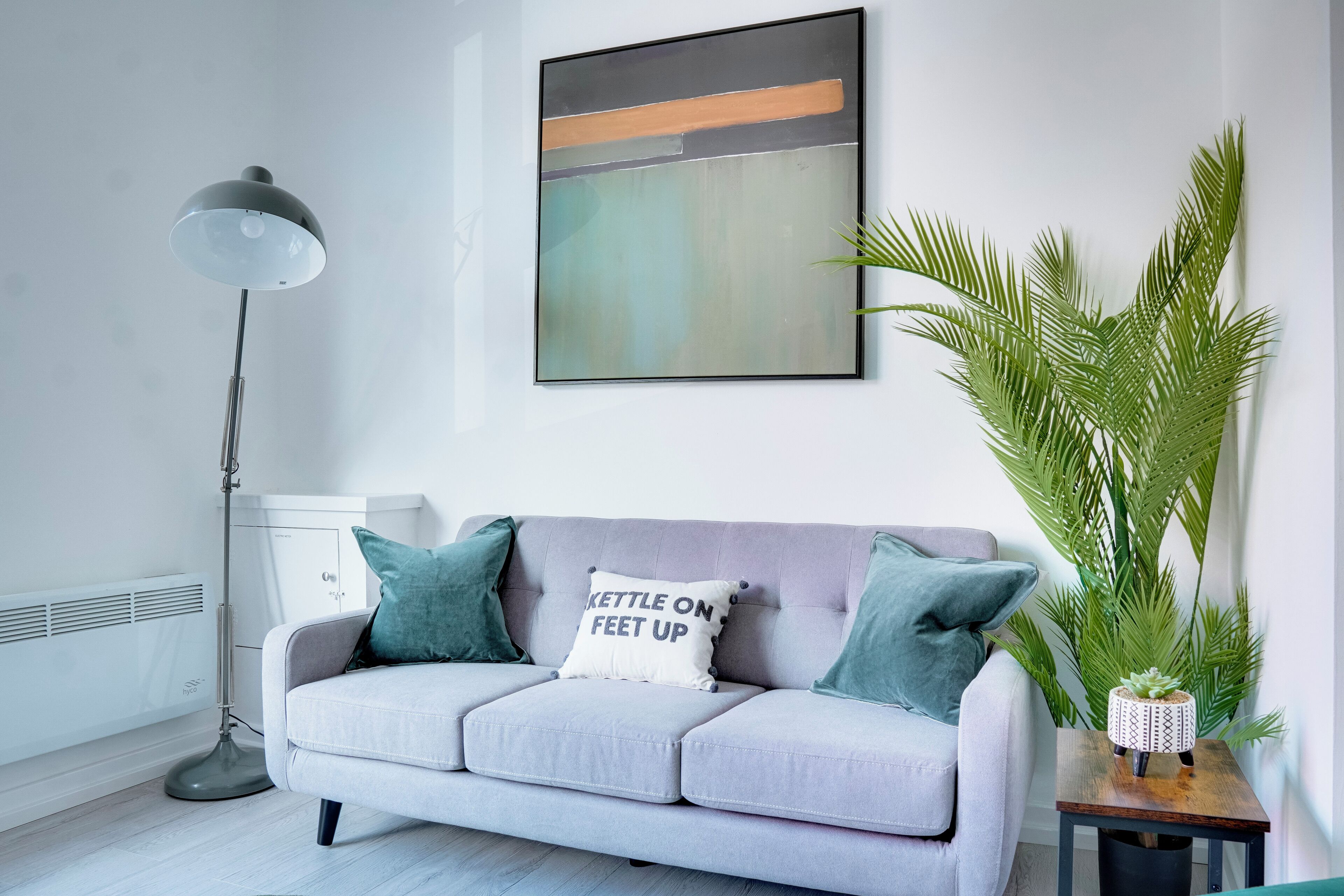 Ein modernes Wohnzimmer mit grauem Sofa, blaustichigen Kissen, einer Stehlampe, einem an der Wand befestigten abstrakten Gemälde und einer Topfpflanze.