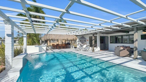 Een zwembad met een pergola, ligbedden en een rieten gebouw op de achtergrond.