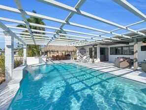 A swimming pool with a pergola overhead, lounge chairs, and a thatched-roof structure in the background.