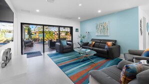 Een moderne woonkamer met een grote glazen deur die leidt naar een terras, een leren zwarte bank, een grijze bank en een kleurrijk gestreept vloerkleed.