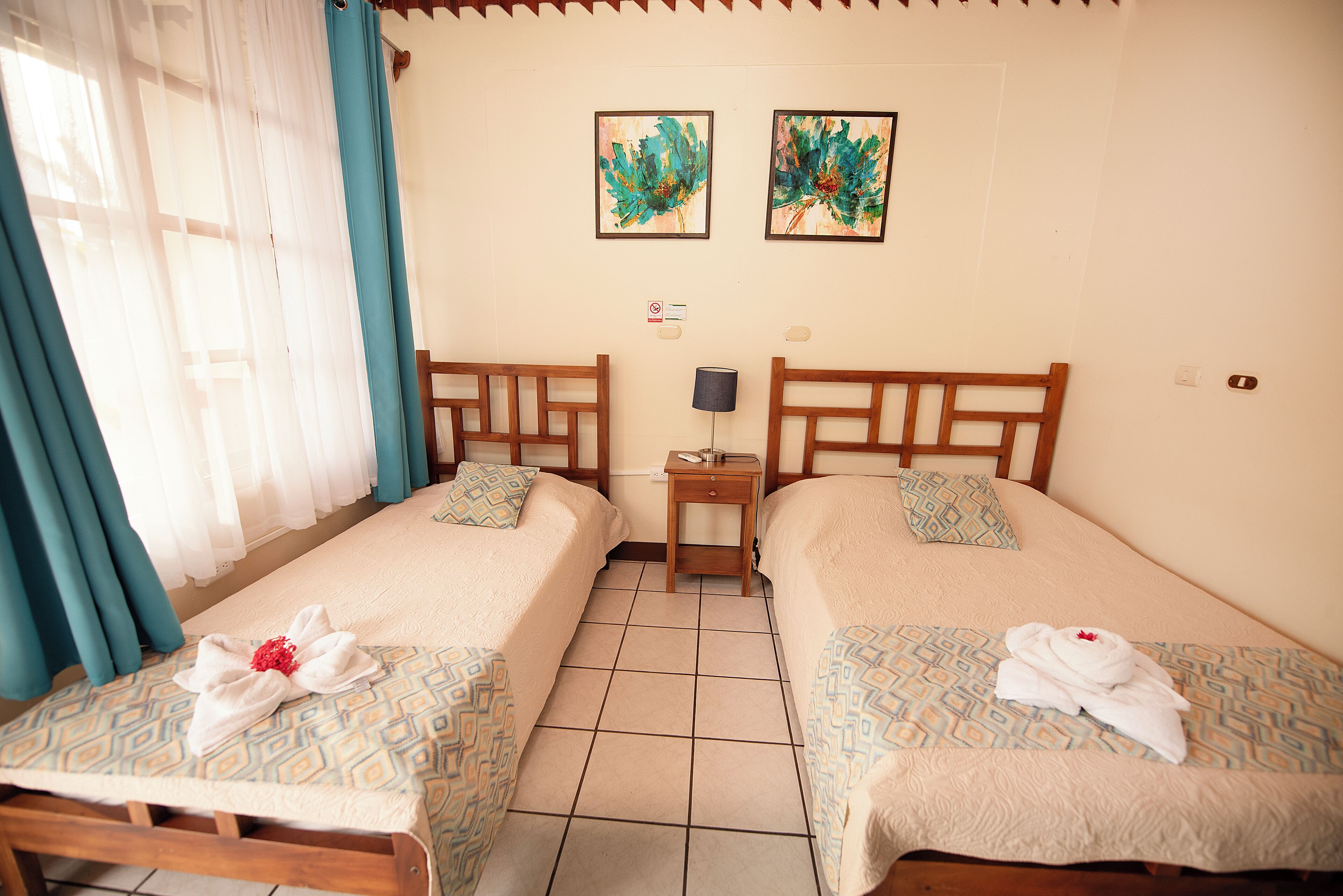 Habitación de hotel con dos camas individuales, una mesita de noche, una lámpara y dos cuadros colgados en la pared.