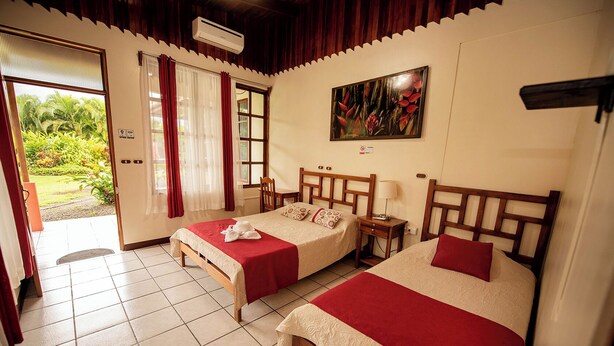 A hotel room with two beds, a window with curtains, a painting on the wall, and an air conditioning unit. - Befox Adventure (La Fortuna)