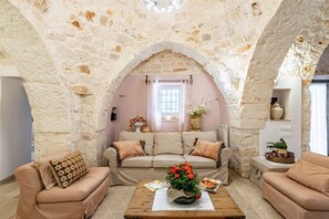 A cozy living room with a stone archway, a sofa set, a coffee table with a flower vase, and a window with curtains.