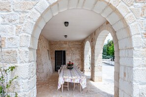 A stone-walled outdoor dining area with a table and chairs, a door, and a lamp.