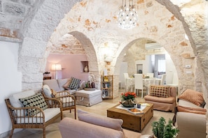 A cozy living room with stone arches, wicker furniture, a coffee table, and a dining area in the background. - Trulli Garden Harmony - Main House by Rentbeat (Martina Franca)