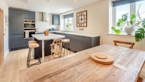 A modern kitchen with grey cabinets, a white countertop, and a wooden dining table with stools. - Station Street Apartment (Cockermouth)
