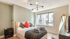 A modern bedroom with a large bed, bedside tables, a mirror, and a window with a city view. - Station Street Apartment (Cockermouth)