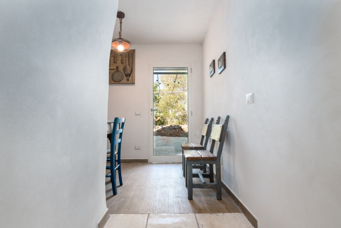 Un couloir avec une porte donnant sur l’extérieur, deux chaises, un luminaire suspendu et un tableau accroché au mur.
