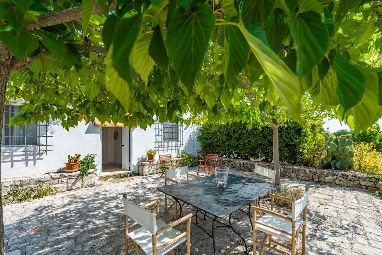 Une terrasse avec une table et des chaises, entourée de plantes en pot et d’un bâtiment blanc doté d’un mur en pierre.
