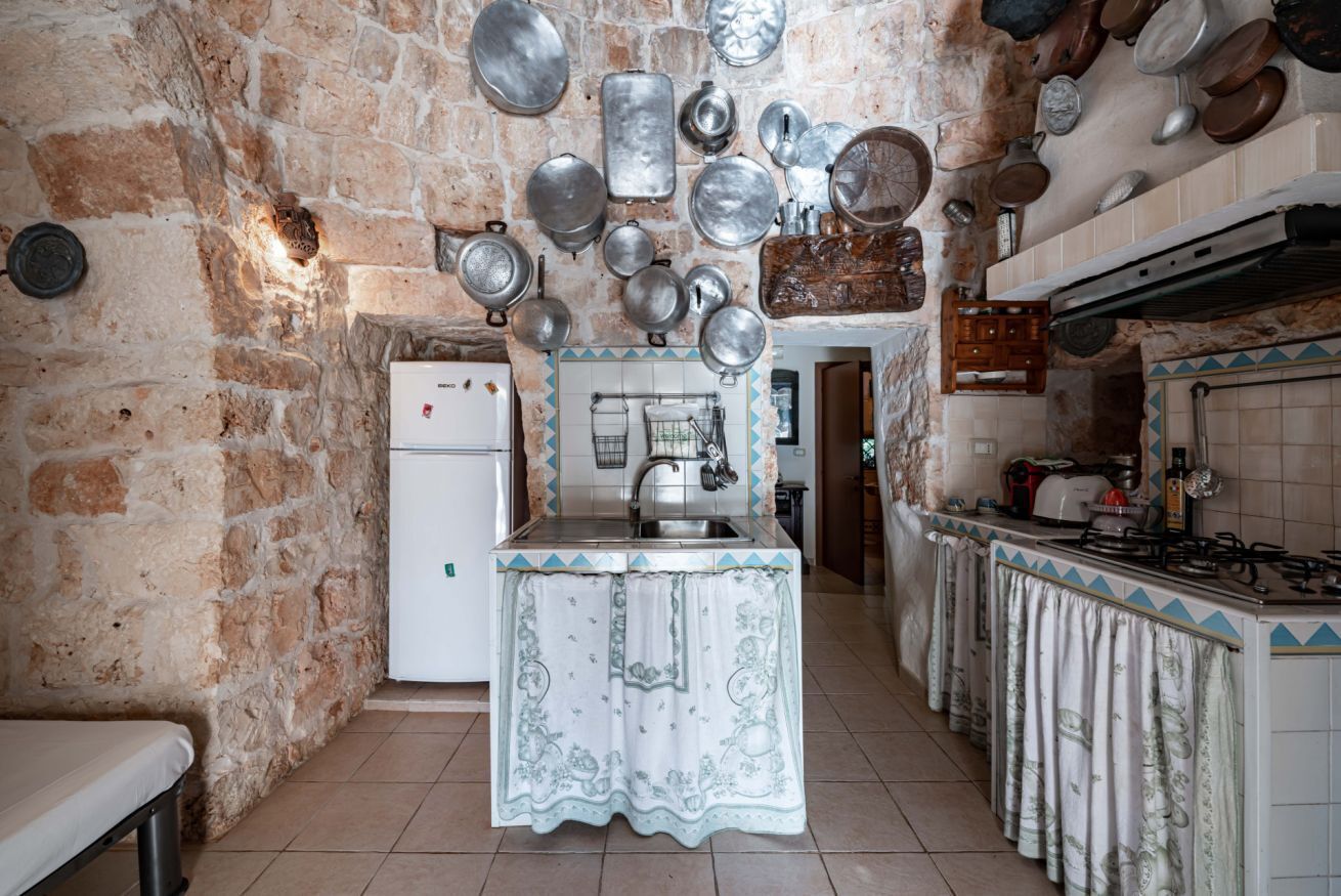 Una cocina con paredes de piedra, un refrigerador, un lavamanos y diversas ollas y sartenes colgando.