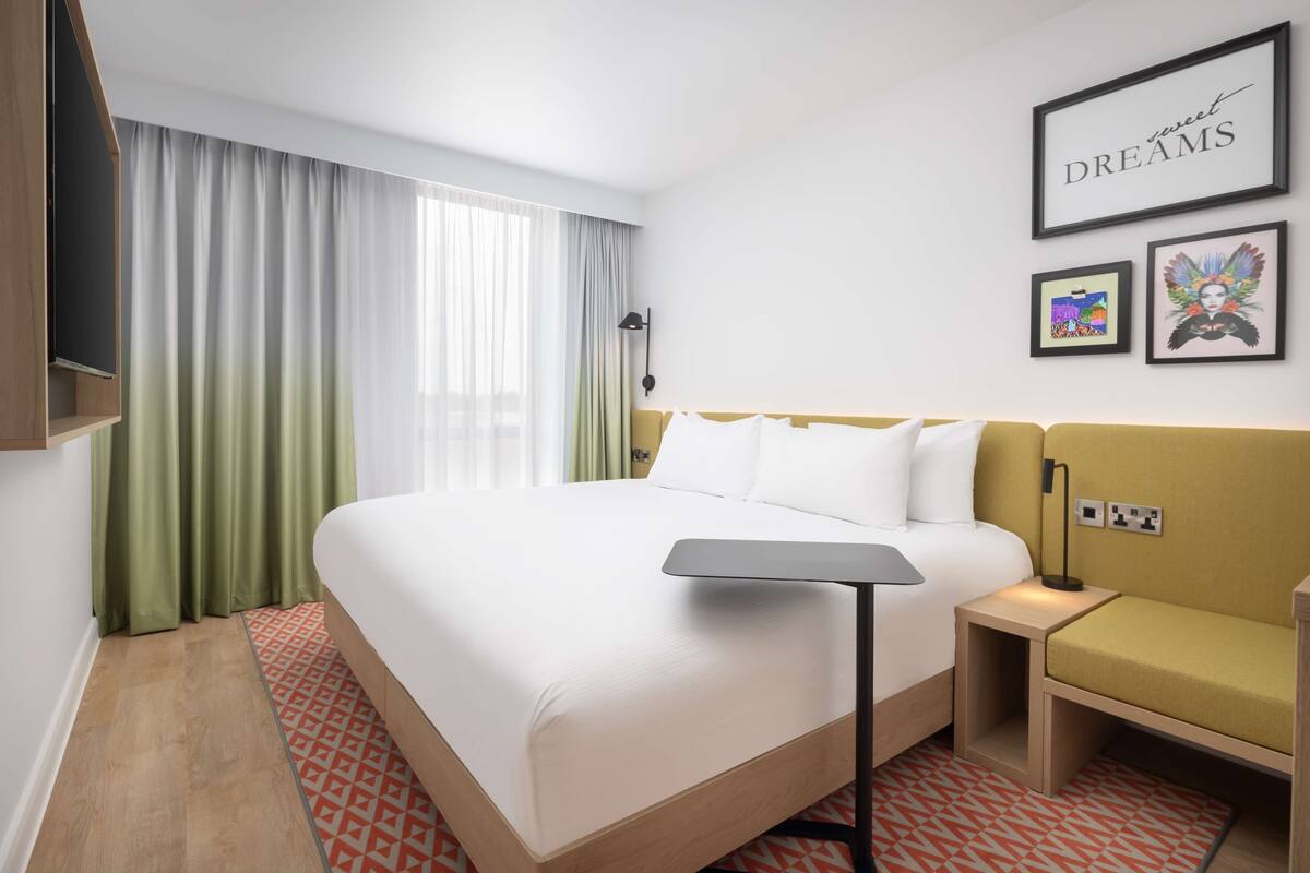 A hotel room with a large bed, a bedside table with a lamp, a small sofa, a TV mounted on the wall, and a window with curtains.