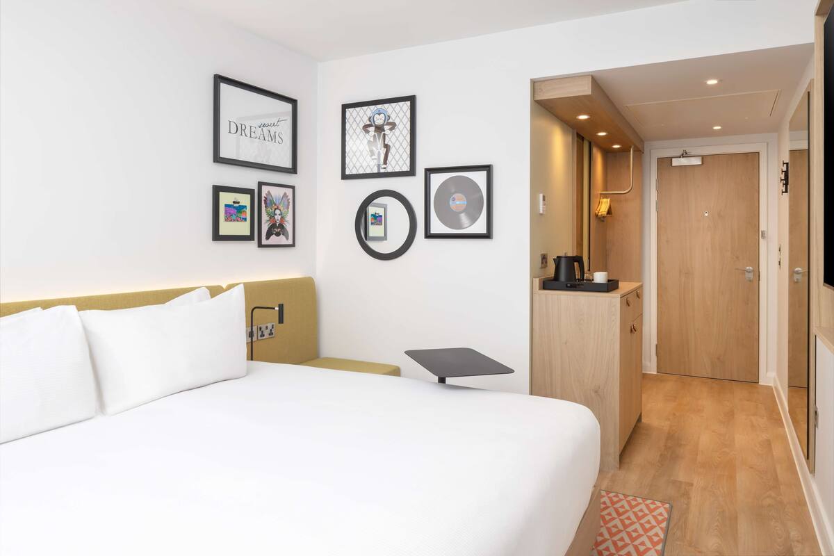 A hotel room with a bed, a bedside table, a desk, and a wall with framed pictures.