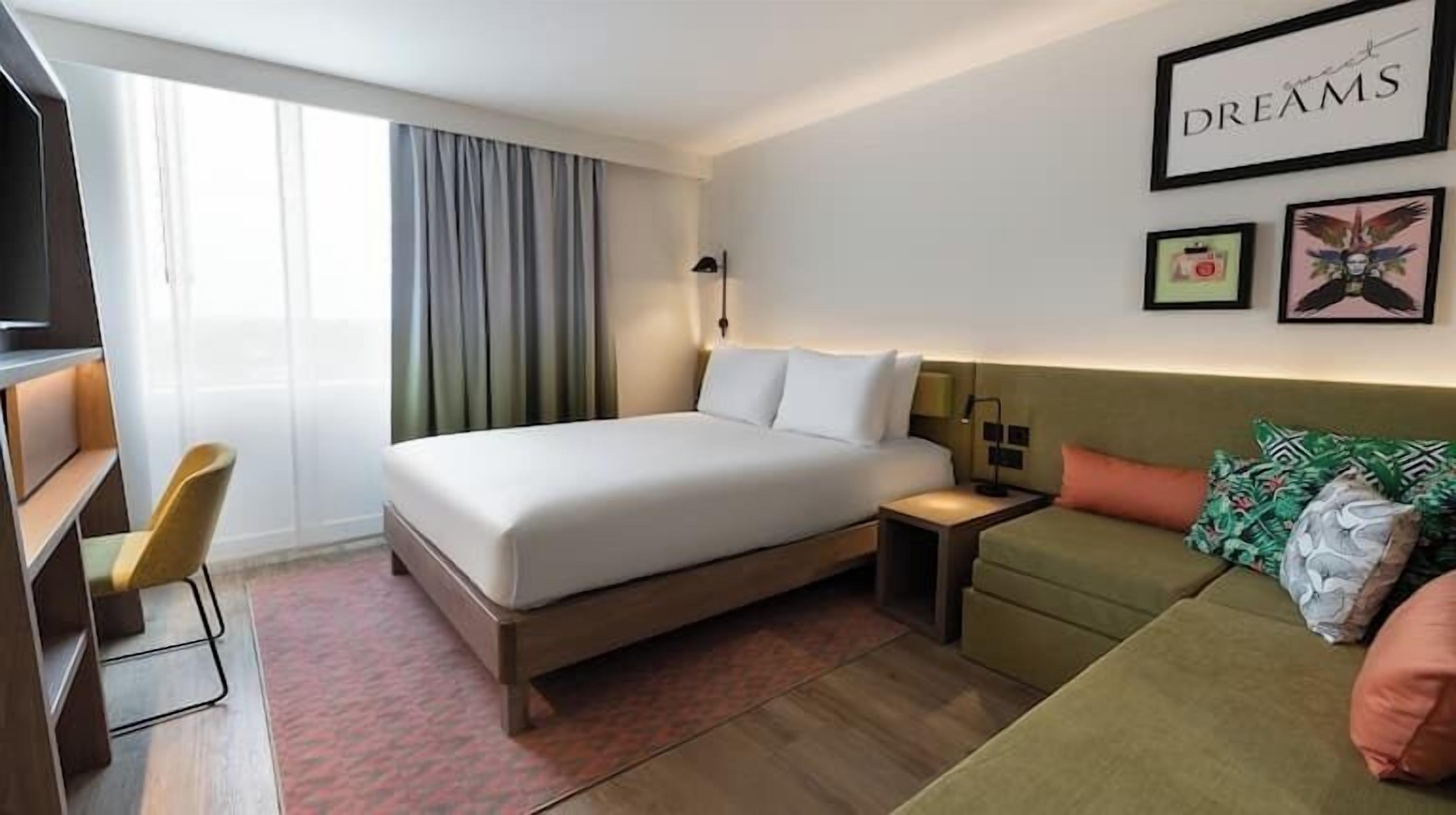 A hotel room with a bed, a desk, a chair, a sofa, a nightstand, and a wall with framed pictures.