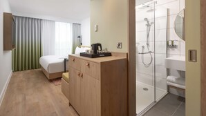 A hotel room with a bed, a wooden wardrobe, a desk with a coffee maker, a shower area with a glass door, and a mirror. - Hampton By Hilton Nuneaton (Nuneaton)