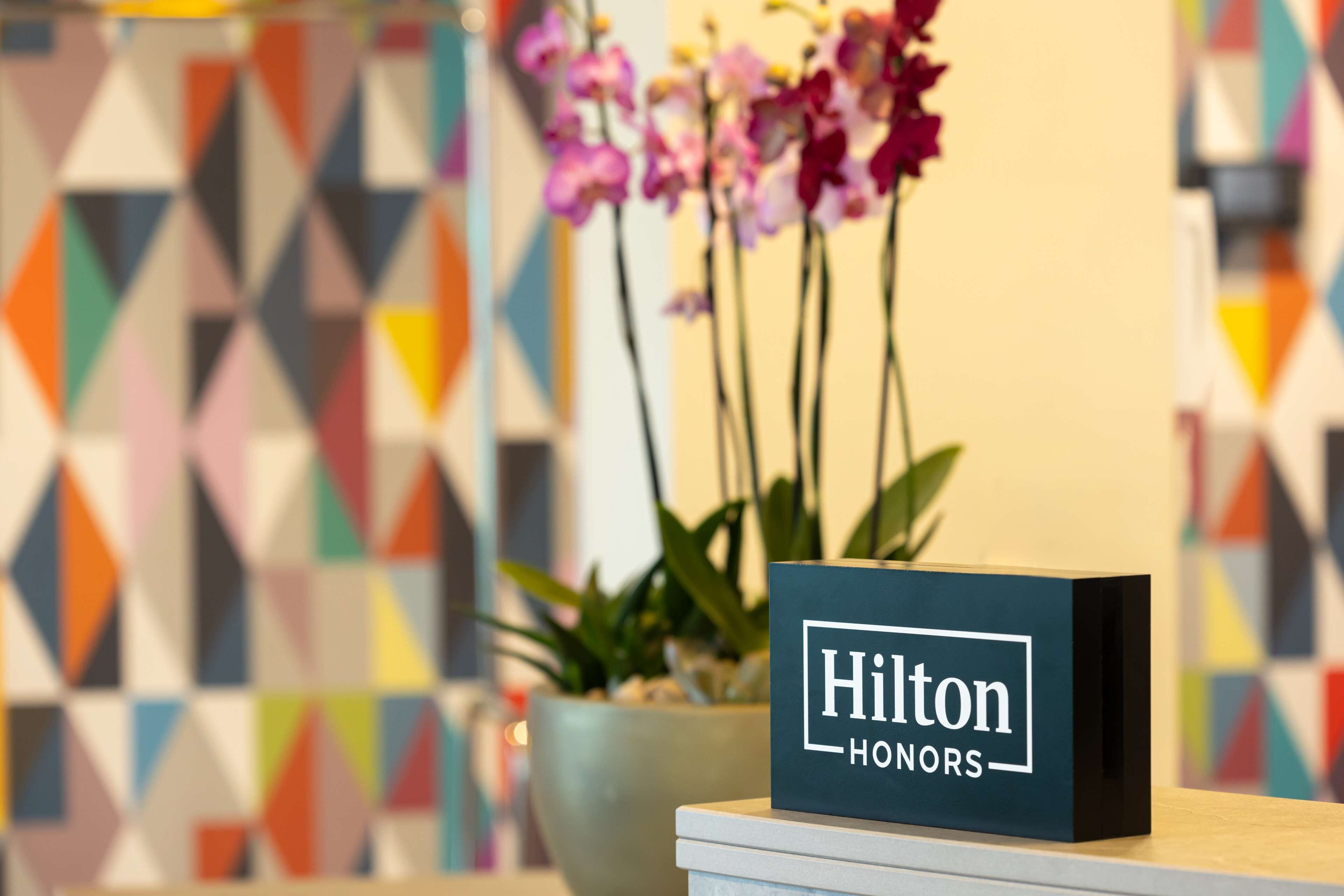 A reception desk with a 'Hilton Honors' sign, a potted plant, and a colorful geometric wall design.