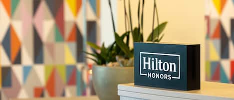 Un comptoir d’accueil orné d’une enseigne « Hilton Honors », d’une plante en pot et d’un motif géométrique coloré sur le mur.