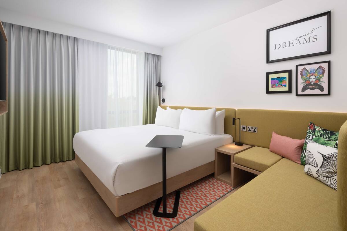 A hotel room with a bed, a bedside table, a nightstand, a sofa, a lamp, and framed pictures on the wall.
