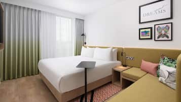 A hotel room with a bed, a bedside table, a nightstand, a sofa, a lamp, and framed pictures on the wall.