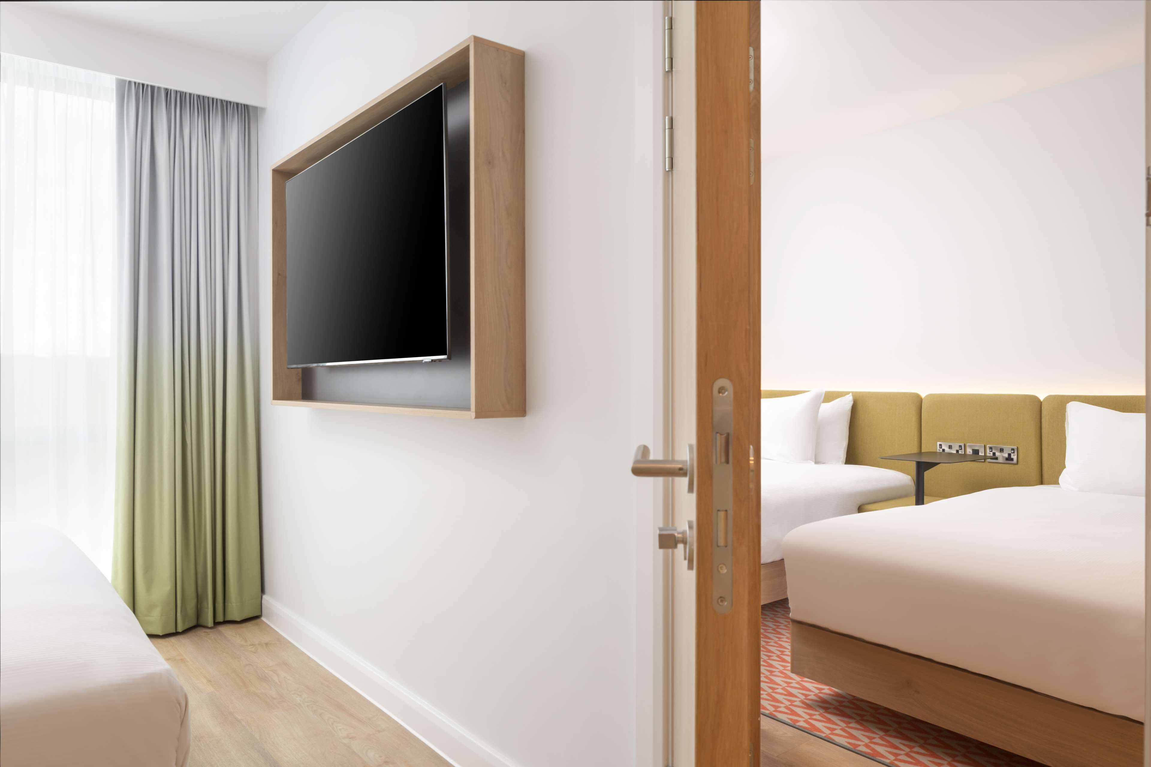 Habitación de hotel con cama, televisión montada en la pared, una mesita y una ventana con cortinas.
