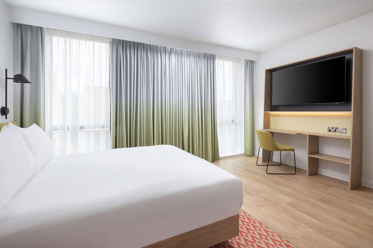 A hotel room with a large bed, a desk with a flat-screen TV, a chair, and a window with curtains.