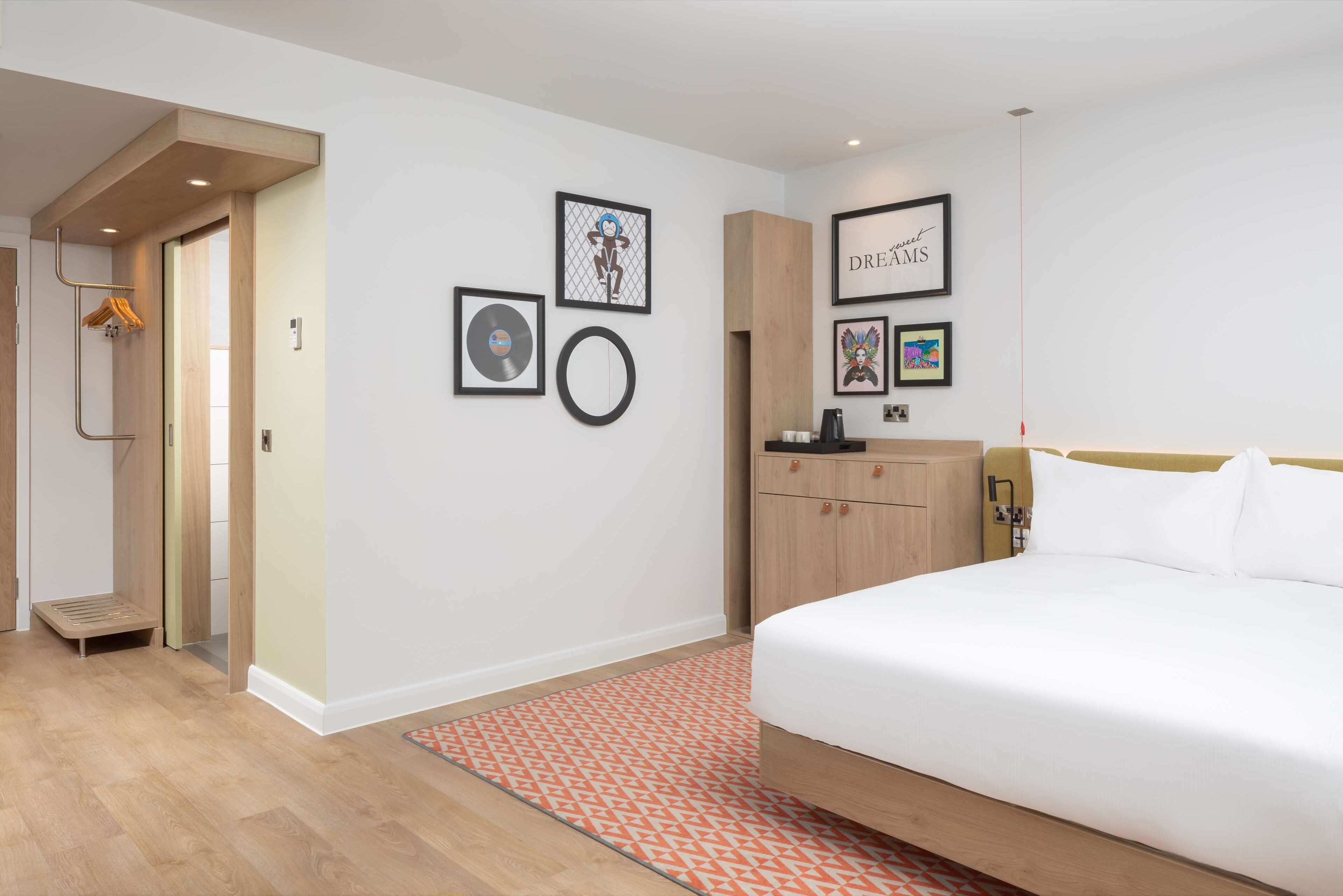 A modern hotel room with a large bed, a wooden wardrobe, a small desk, and a wall with framed pictures.