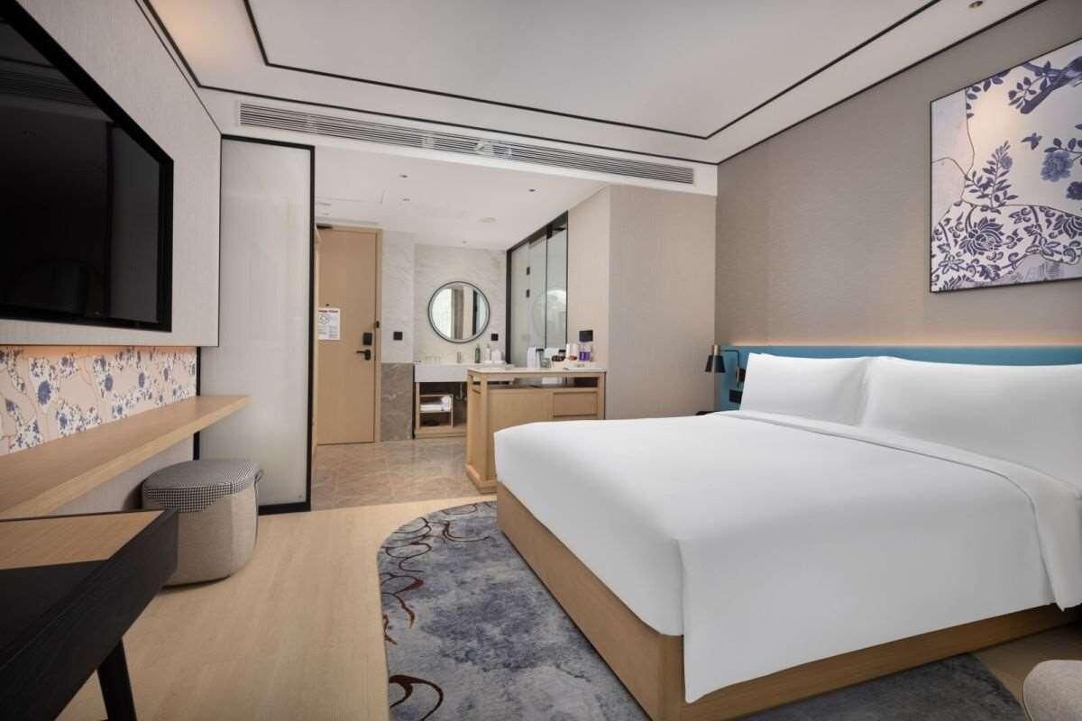 A modern hotel room with a large bed, a desk, a TV, a bathroom with a mirror, and a decorative wall piece.