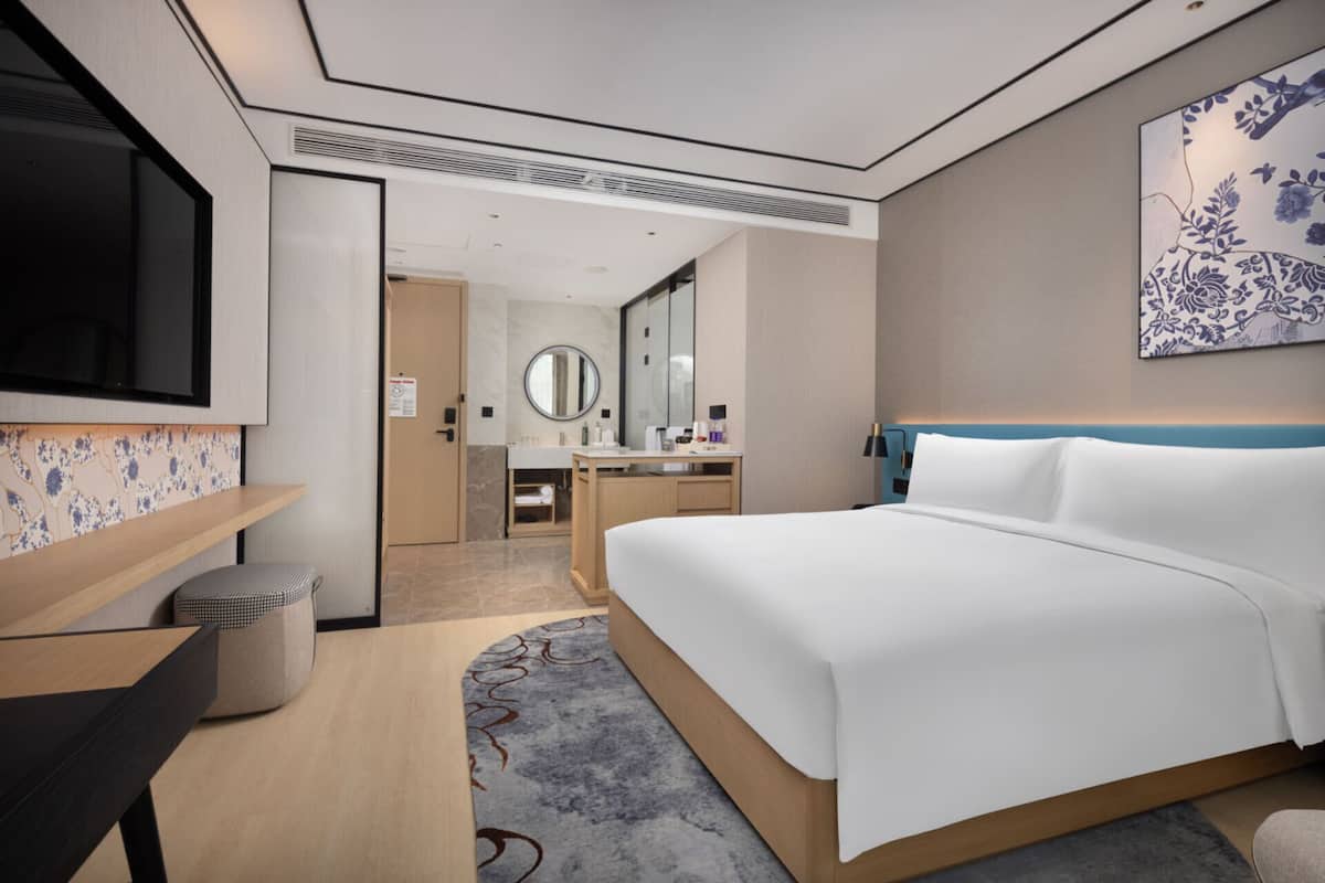 Une chambre d’hôtel moderne avec un grand lit, un bureau, une télévision, une salle de bain avec un miroir et un tableau au mur.