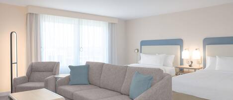 Quarto de hotel com cama, sofá, poltrona, mesa pequena e janela com cortinas.