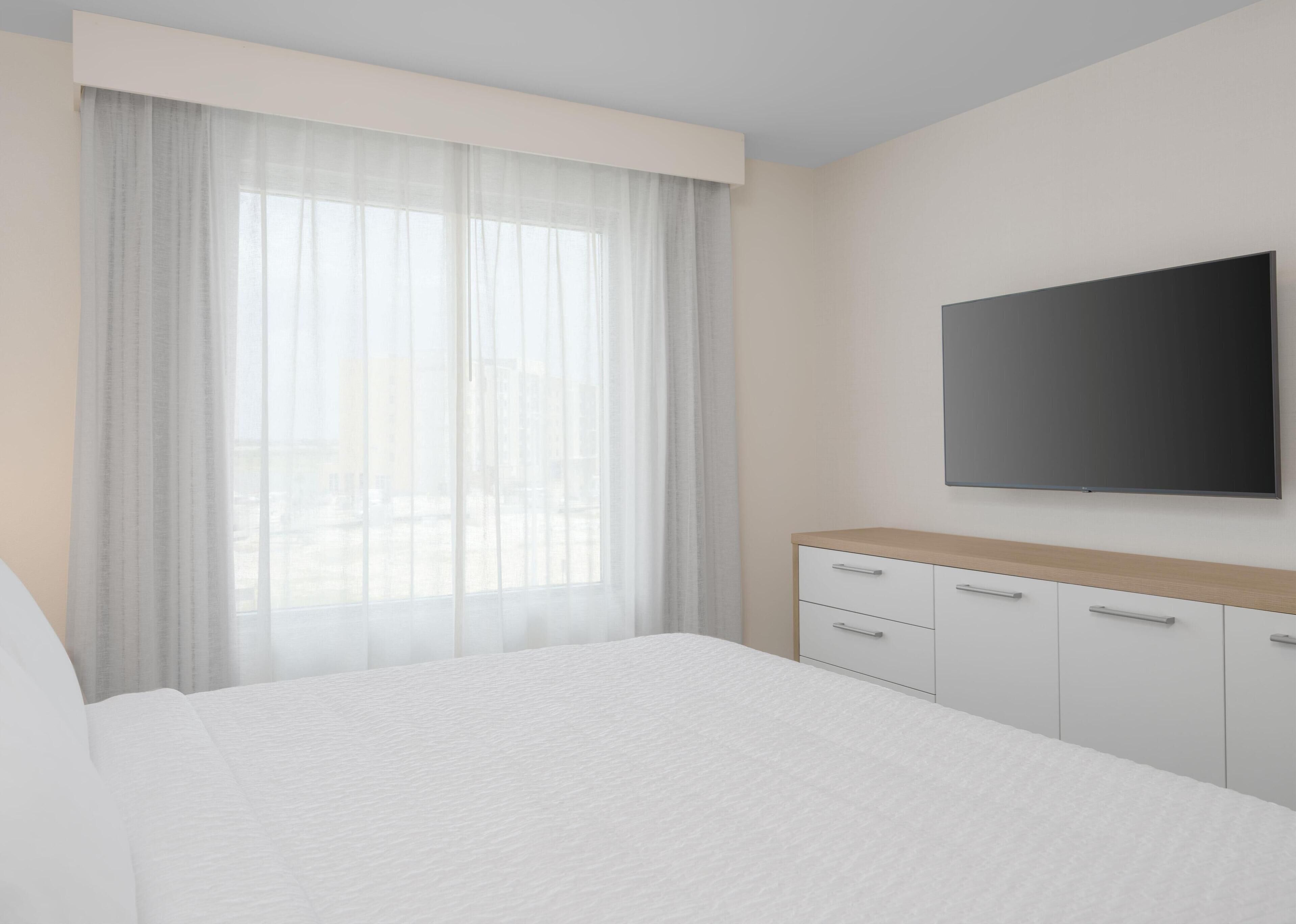 Habitación de hotel con una cama grande, televisor montado en la pared y una ventana con cortinas traslúcidas.