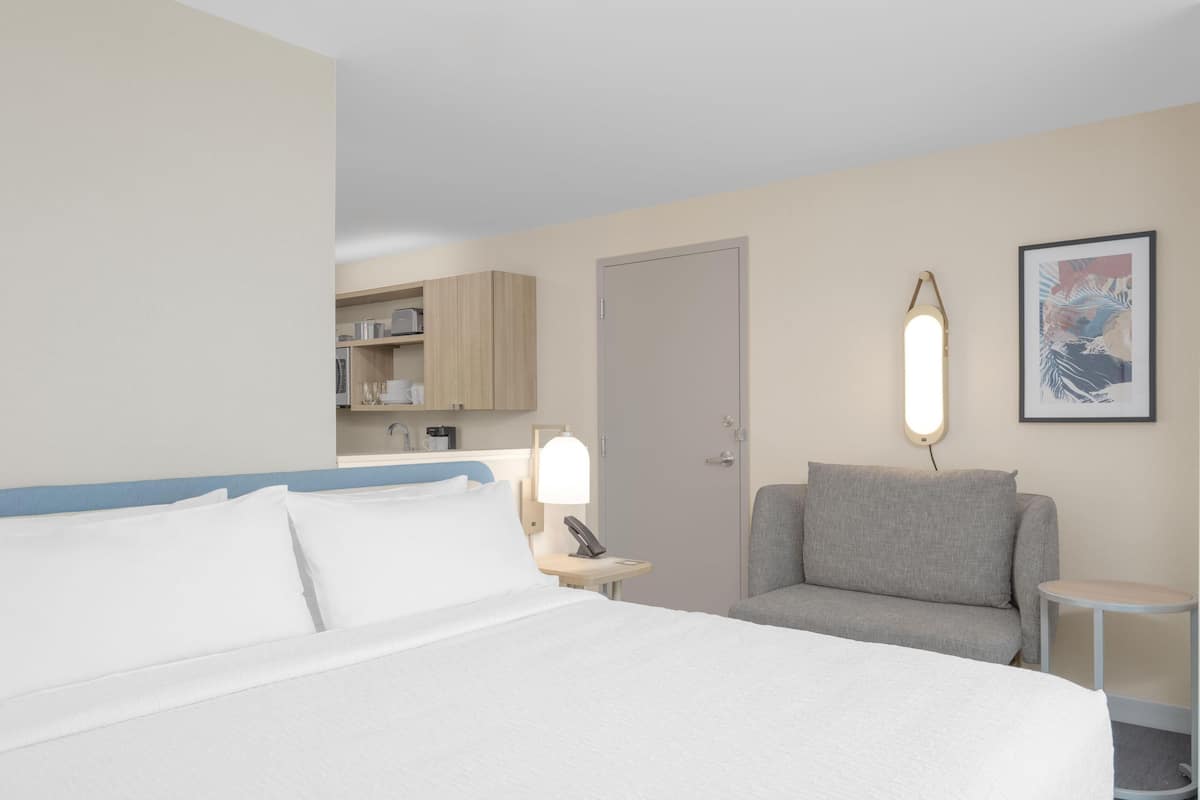 Habitación de hotel con una cama grande, un sofá gris, una mesita, una lámpara de noche, una puerta y un cuadro en la pared.