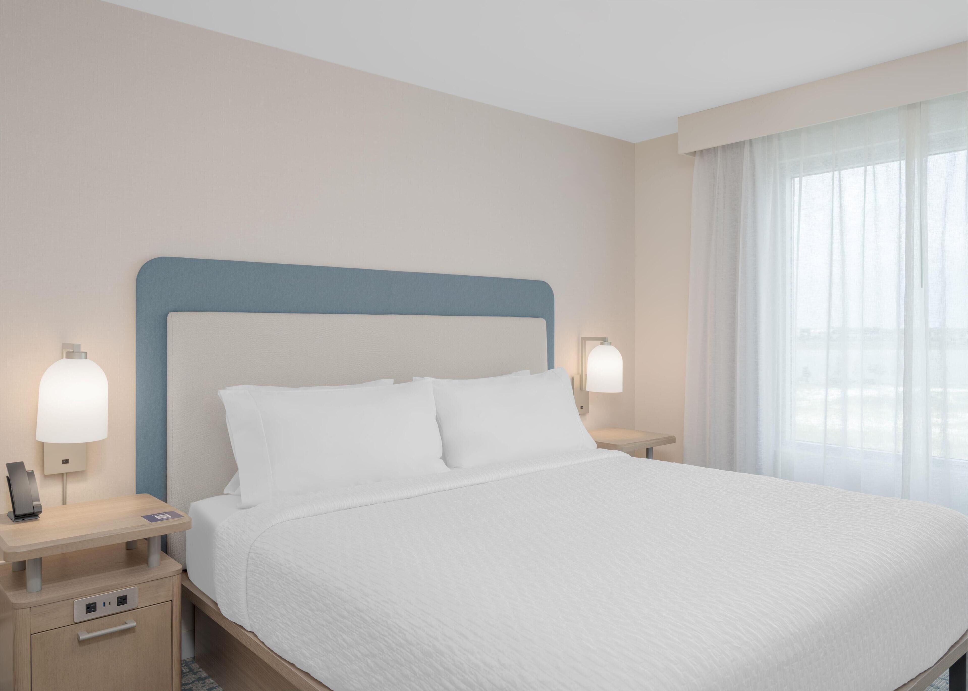 A hotel room with a large bed, two bedside tables, a nightstand, a lamp, and a window with sheer curtains.