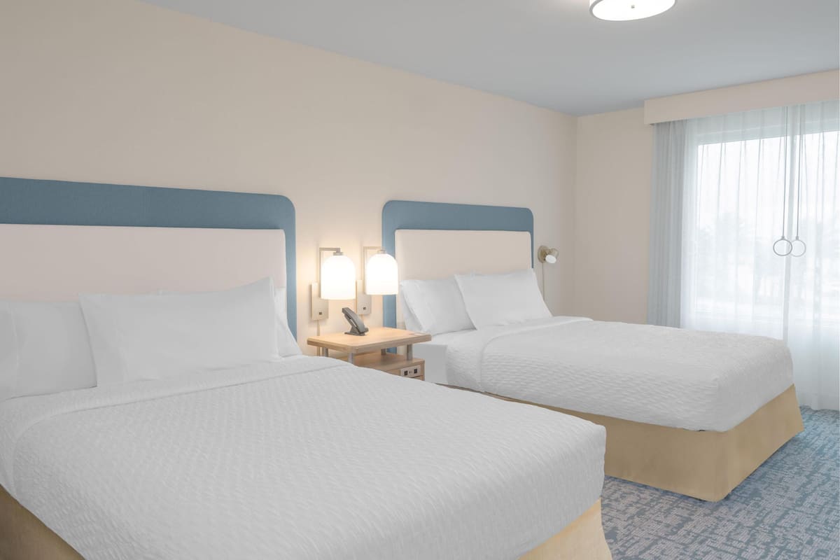 Una habitación de hotel con dos camas, una mesita de noche con lámpara y una ventana con cortinas traslúcidas.