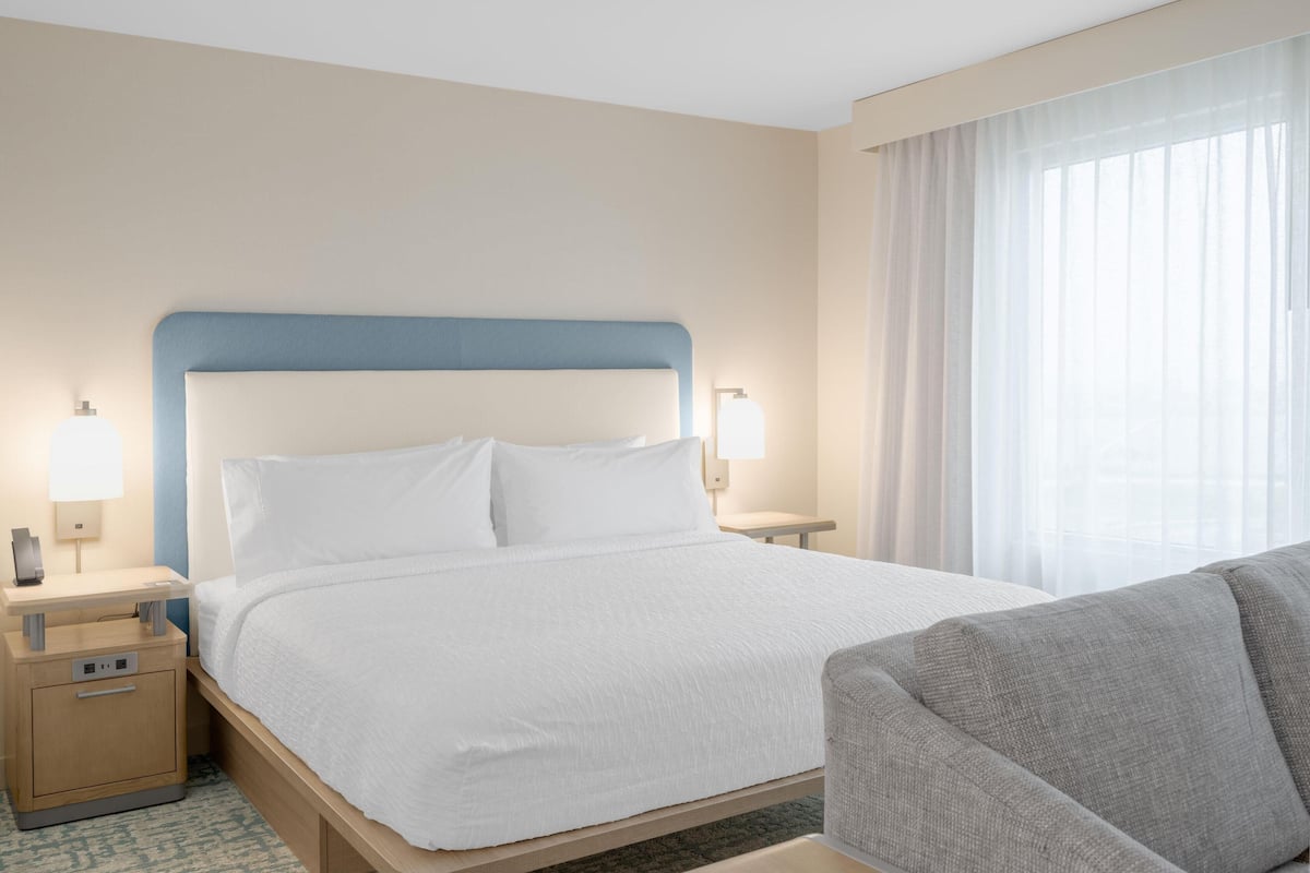 Una habitación de hotel con una cama grande, una mesita de noche con lámpara, una silla y una ventana con cortinas.