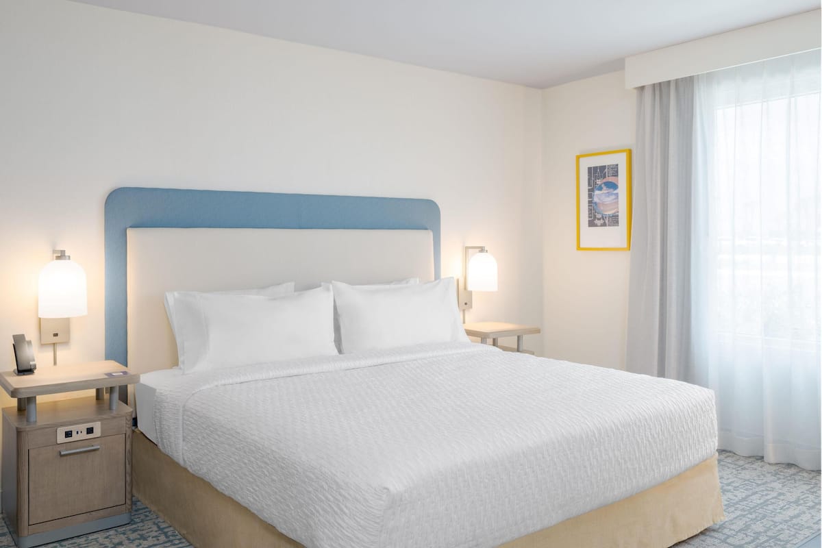 Una cama bien hecha con cabecera azul, dos lámparas de noche, una mesita y un cuadro en la pared.