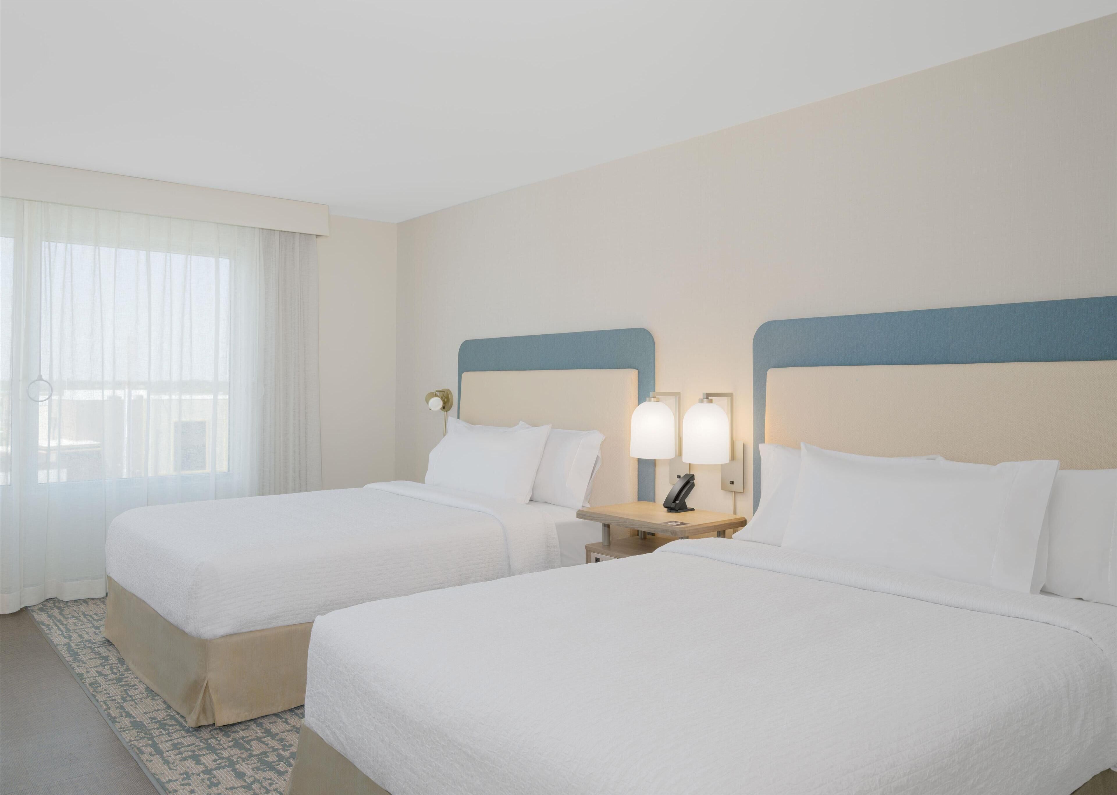 Una habitación de hotel con dos camas, una mesita de noche con lámpara y una ventana con cortinas traslúcidas.