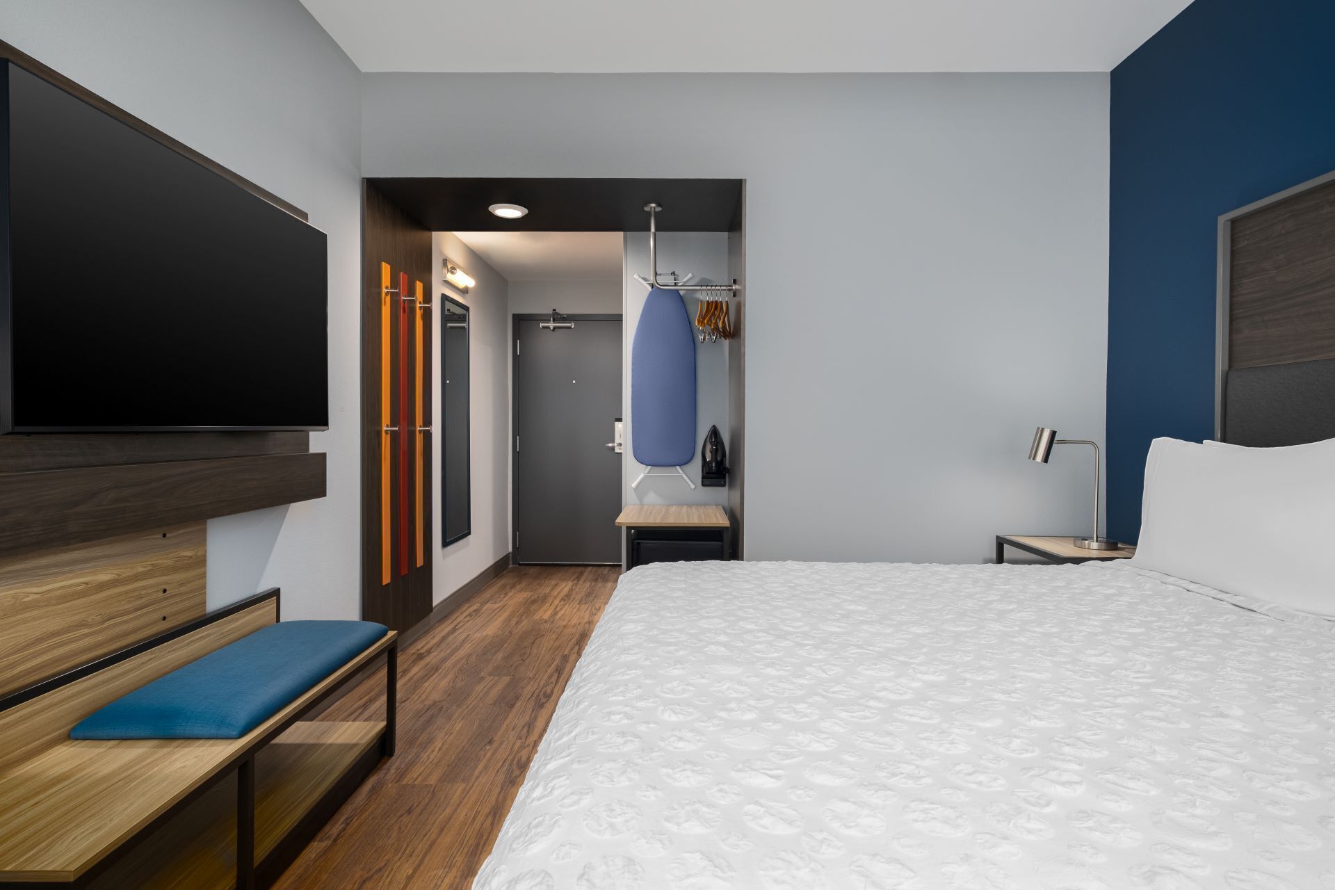 Une chambre d’hôtel comprenant un lit, un bureau, une banquette, une télévision, une armoire, un miroir et une porte donnant accès à une salle de bain.
