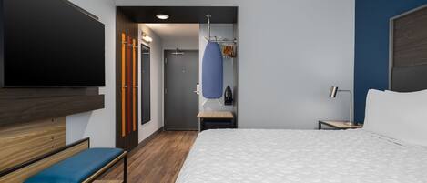 A hotel room with a bed, a desk, a bench, a TV, a wardrobe, a mirror, and a door leading to a bathroom.