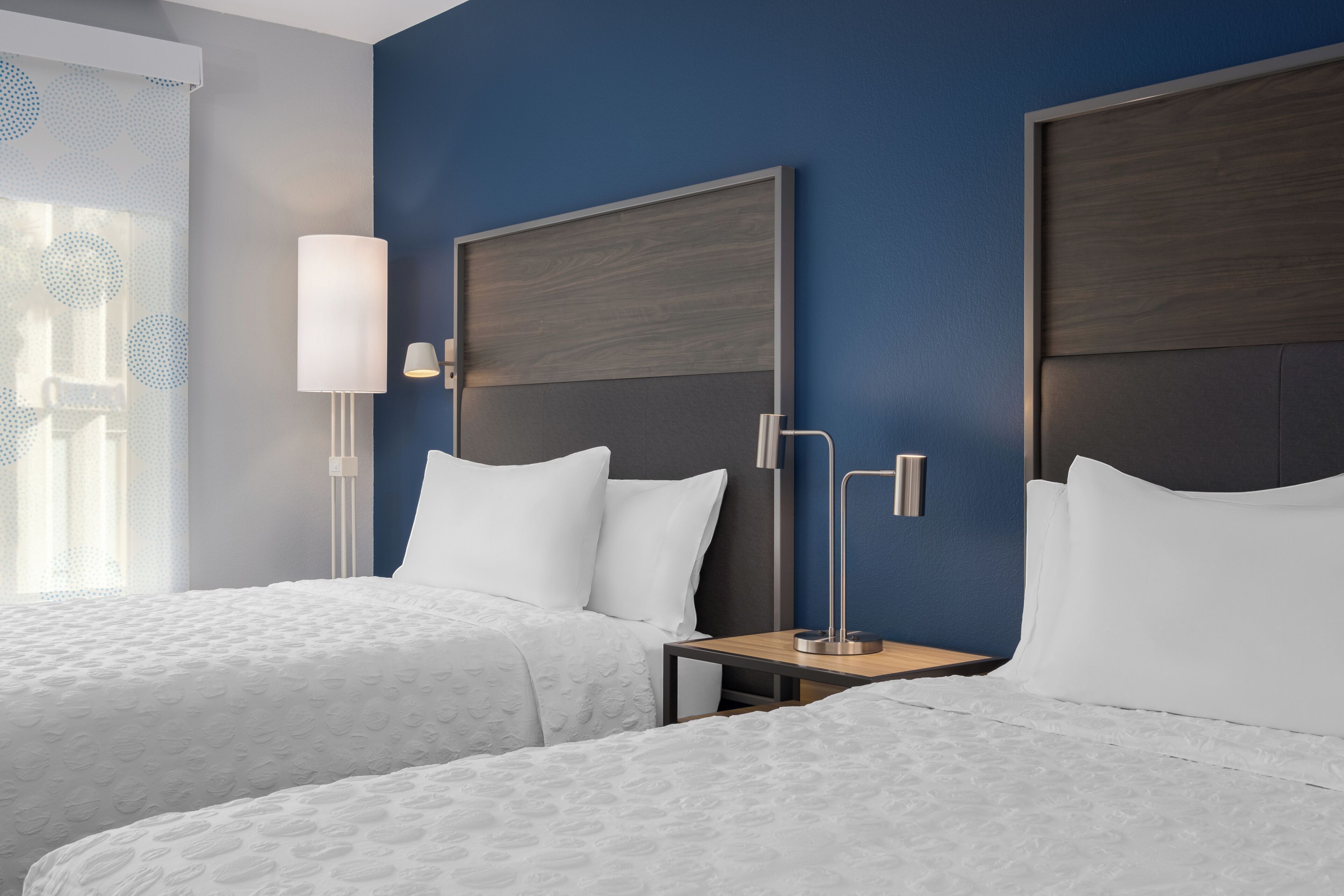 A hotel room with two beds, a nightstand, a lamp, and a wall-mounted headboard.