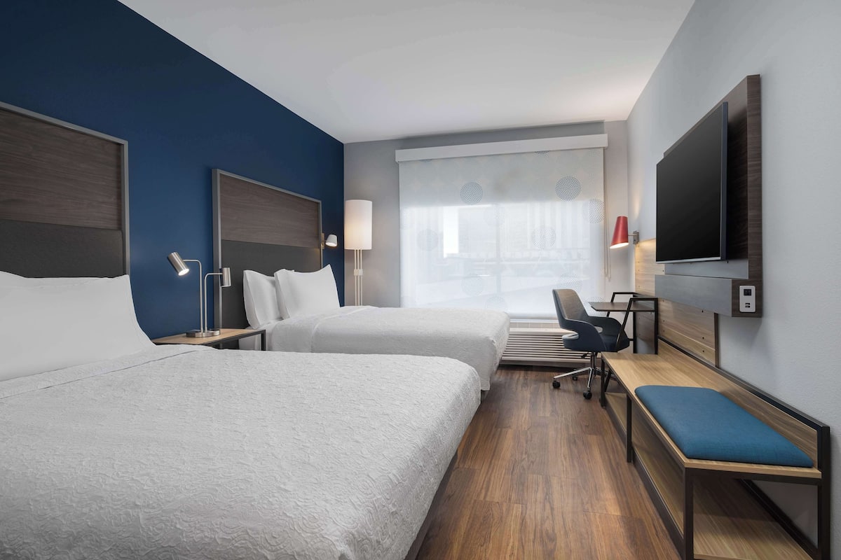 A hotel room with two beds, a desk, a chair, a TV, and a window with curtains.