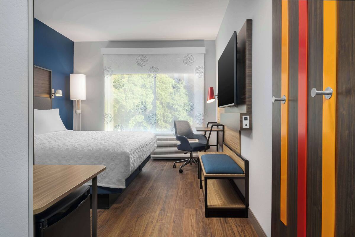 A hotel room with a bed, desk, chair, TV, and a window with a view of trees.