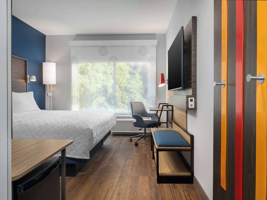 Habitación de hotel con cama, escritorio, silla, televisor y ventana con vistas a árboles.