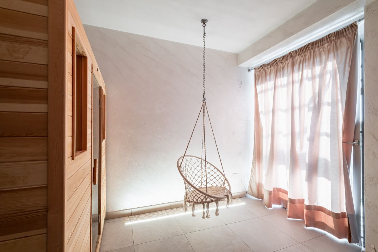 Une pièce dotée d’un fauteuil suspendu, de murs recouverts de panneaux en bois, d’une grande fenêtre ornée de rideaux légers et d’un sol carrelé.