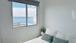 Un dormitorio con un amplio ventanal con vistas al mar, una cama con ropa de cama blanca y dos almohadas, una mesita de noche con lámpara y vista al océano.
